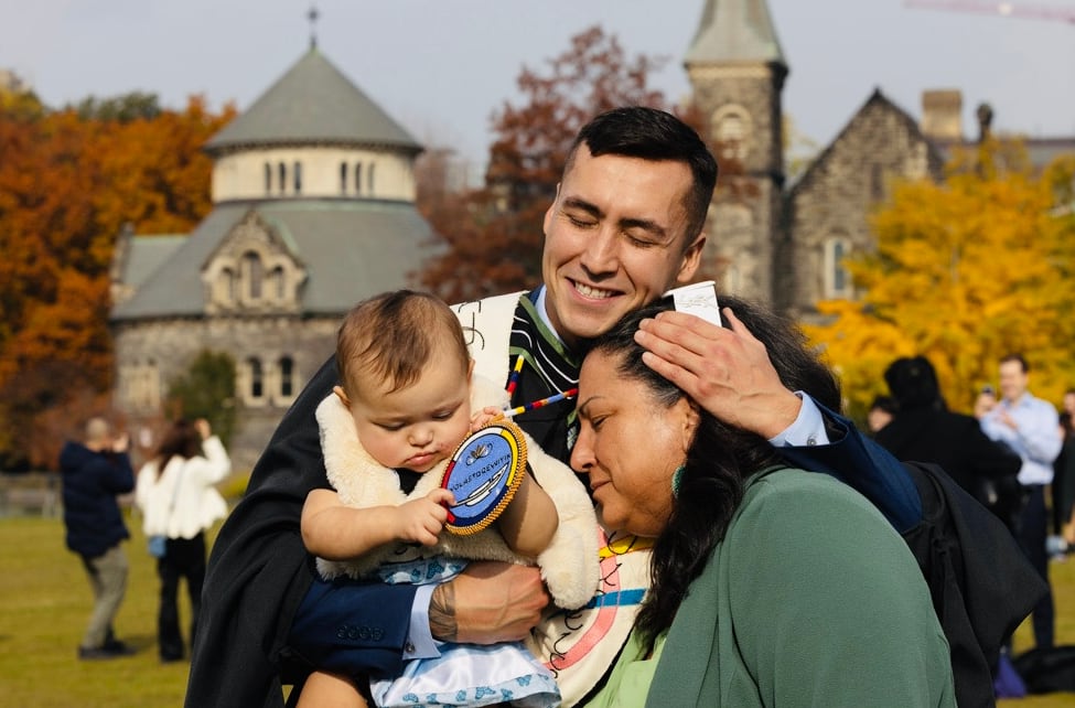 A man in a graduation robe holding a baby and hugging a woman.