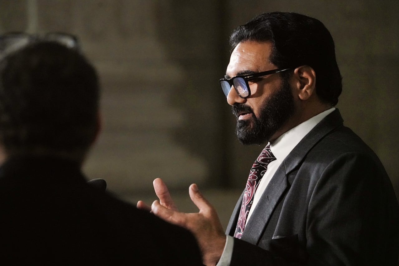 A man with dark hair and a dark beard, wearing a light striped suit and tie, speaks during a government legislative session.
