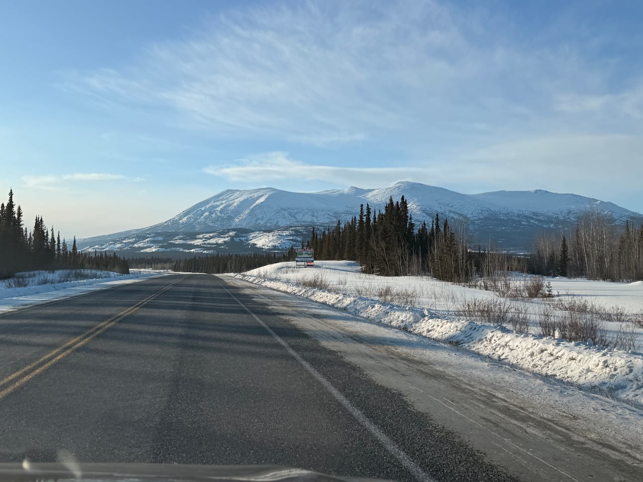 A photo taken from the front seat of a car looking at the road ahead with mountains and trees in the background. 