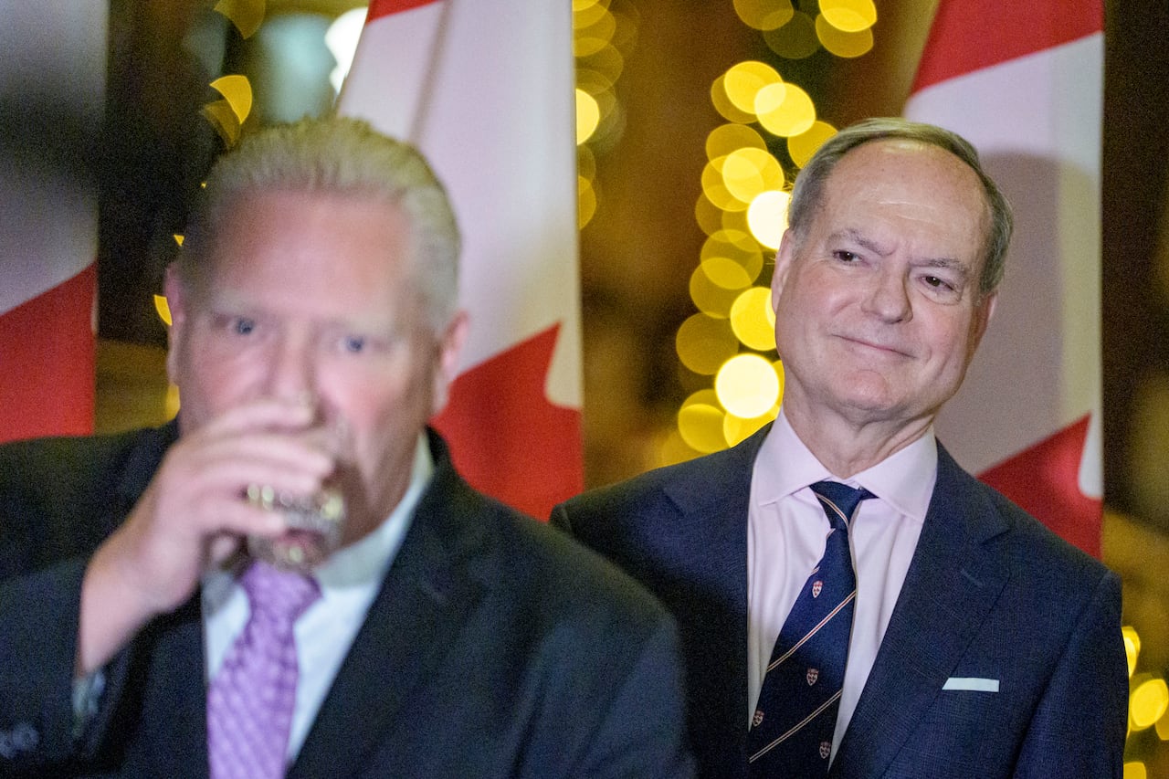 A man takes a drink of water while another looks over his shoulder, smiling.