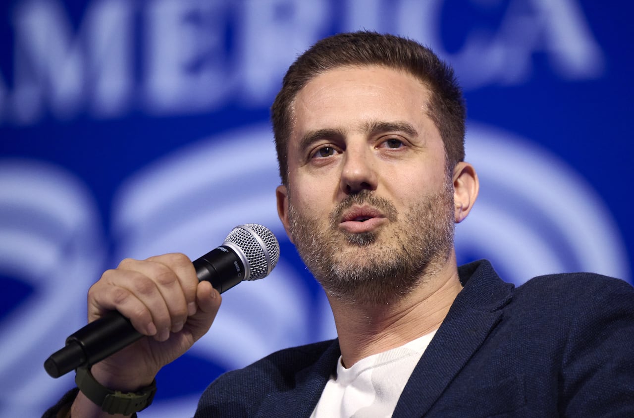 A man in a navy blue suit jacket is pictured holding a handheld microphone.