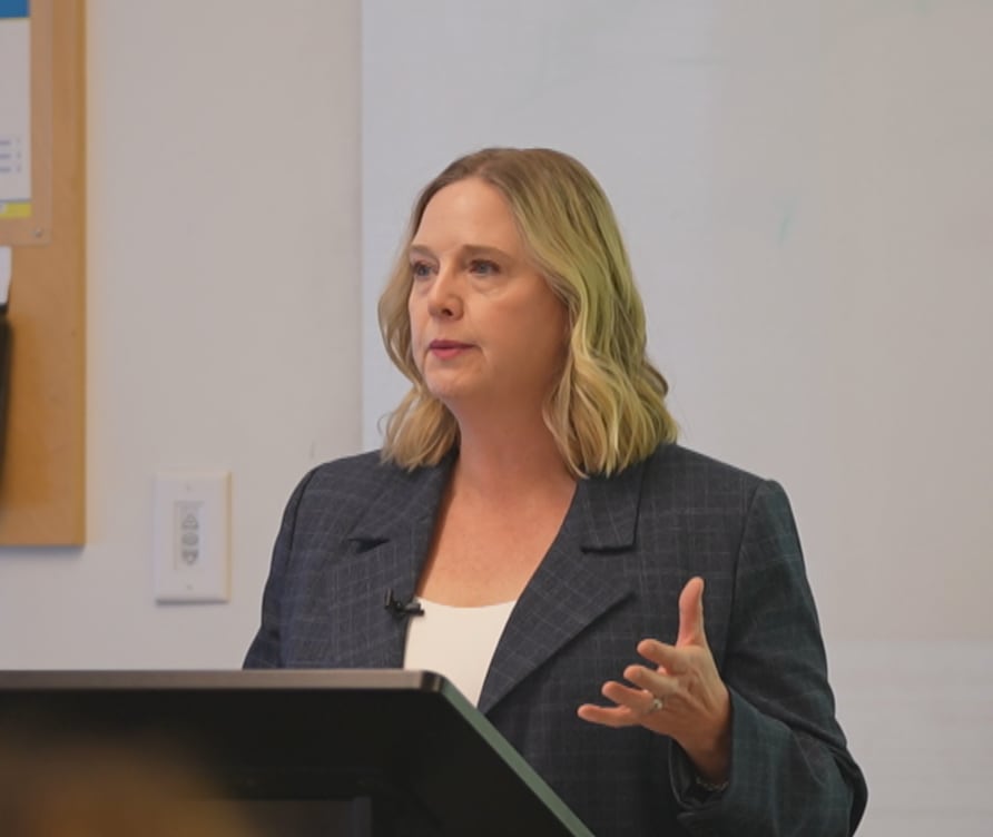 A woman stands behind a podium at the front of a university lecture hall.
