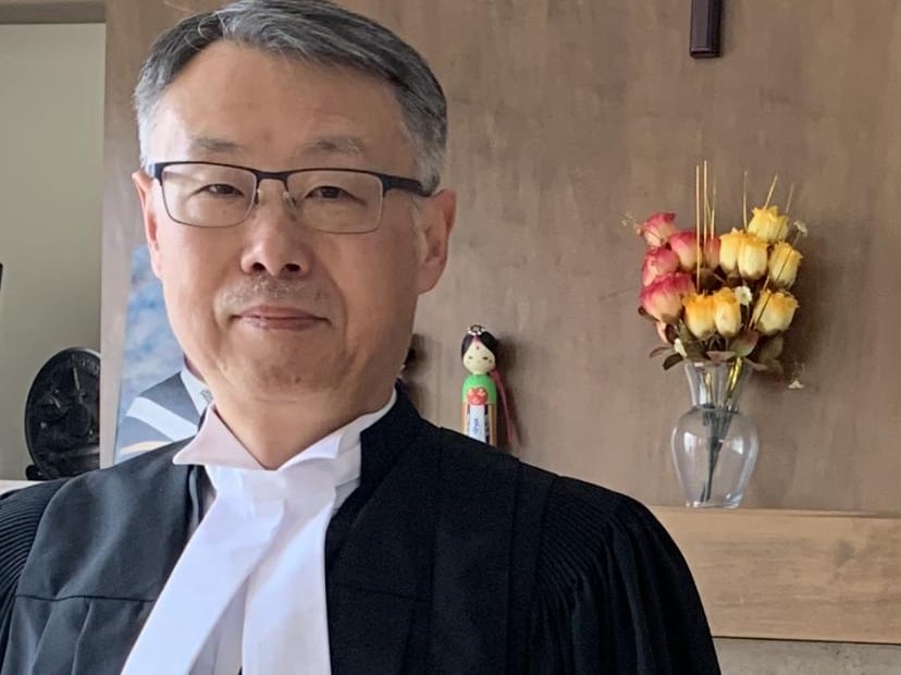 A male lawyer in stands in his robes in front of a crucifix.