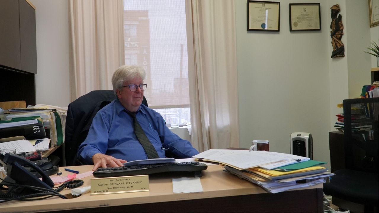 A man with white hair, glasses and a blue button down shirt sits at a desk.