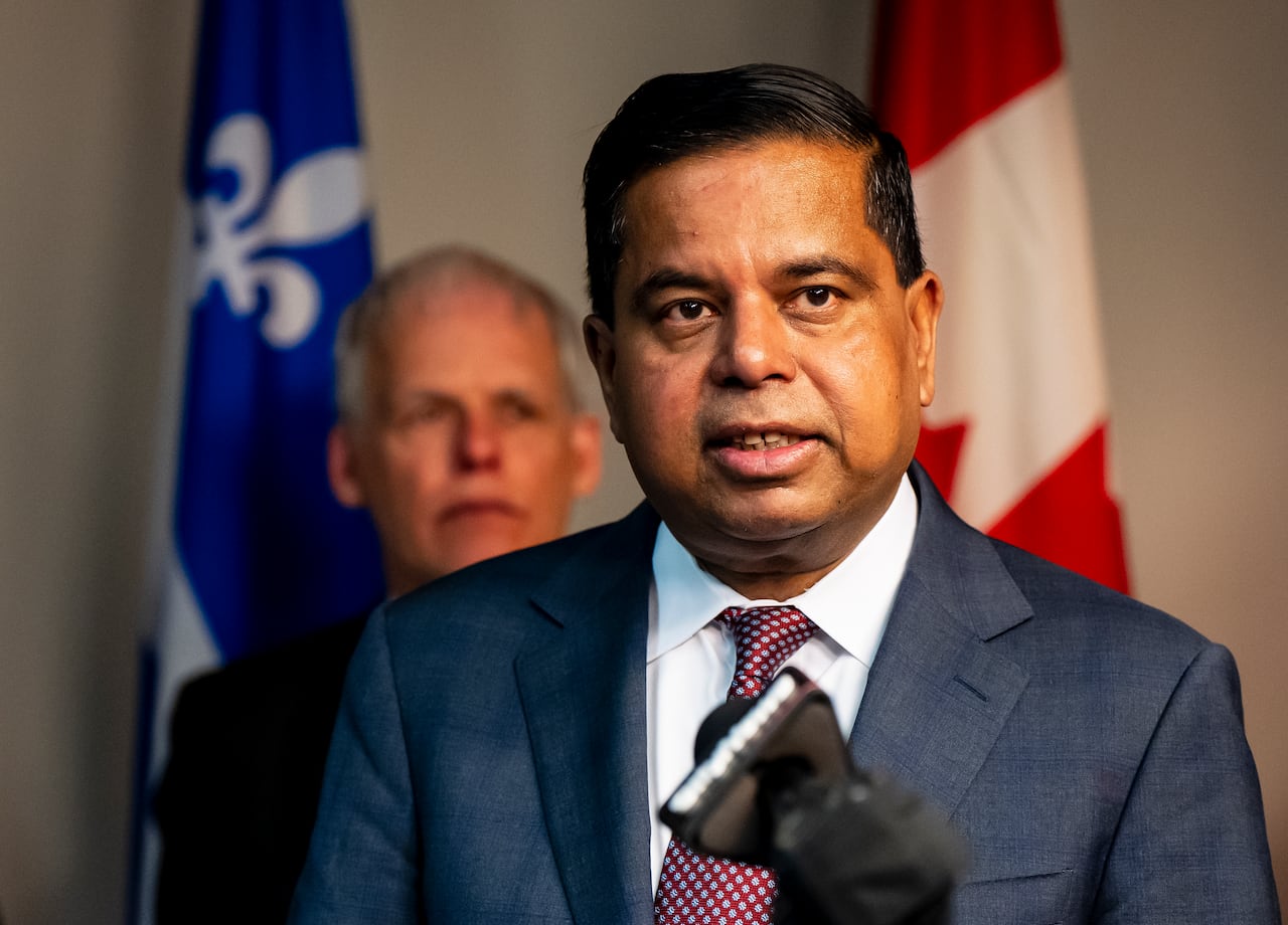 A man speaks in front of another man and a Quebec flag and a Canadian flag.