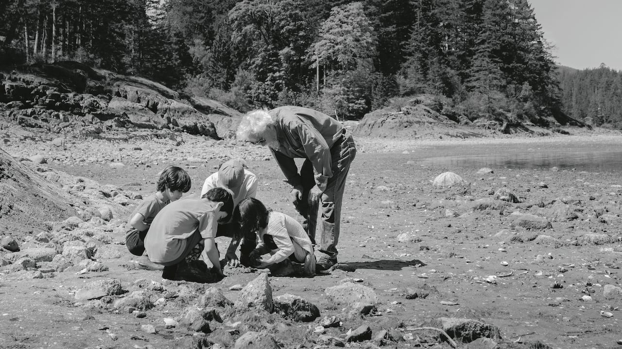 A man with white hair leans over and looks at four kids playing in the beach sand. 