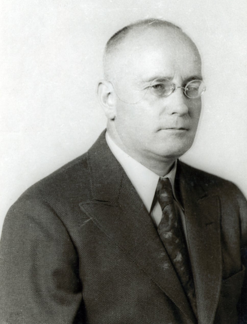 A black-and-white photo shows a man wearing a suit and tie with glasses.
