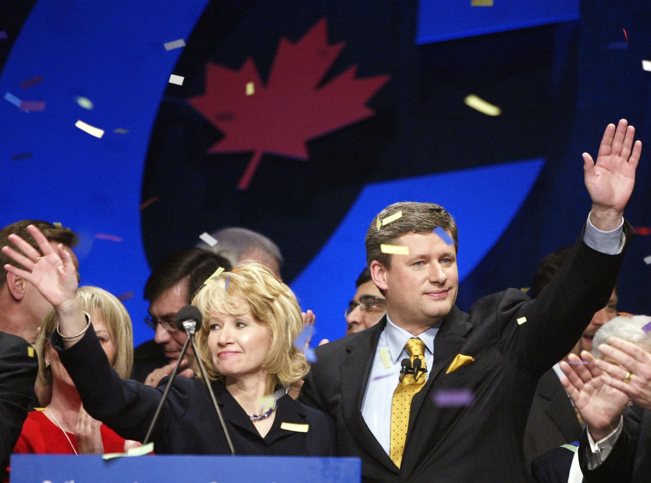 A man and woman wave in front of a blue Conservative sign backdrop, as confetti falls on them