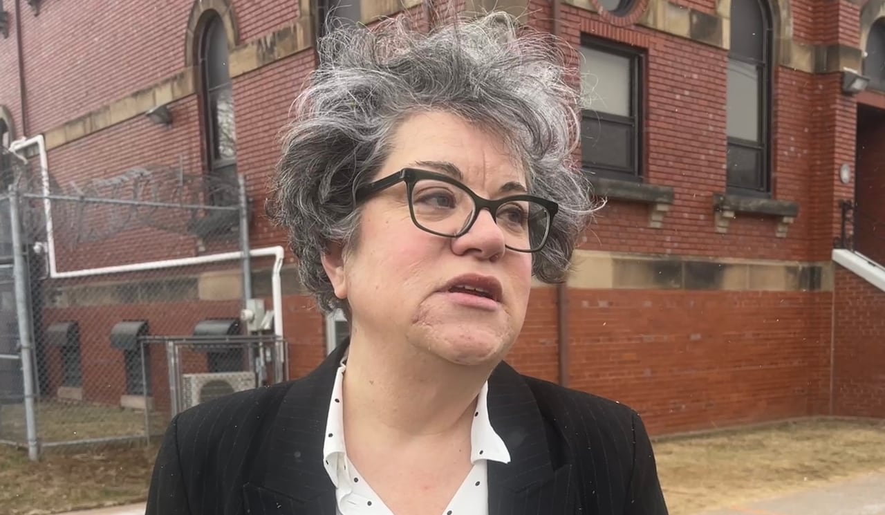 A dark haired woman with glasses stands in front of a microphone with a CBC logo