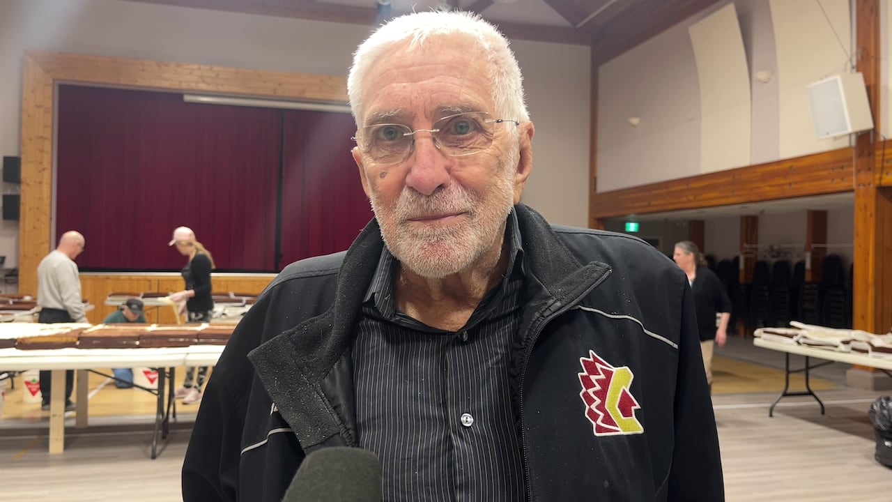 An old man is seen in a large community centre, with people behind him working on a row of cakes.