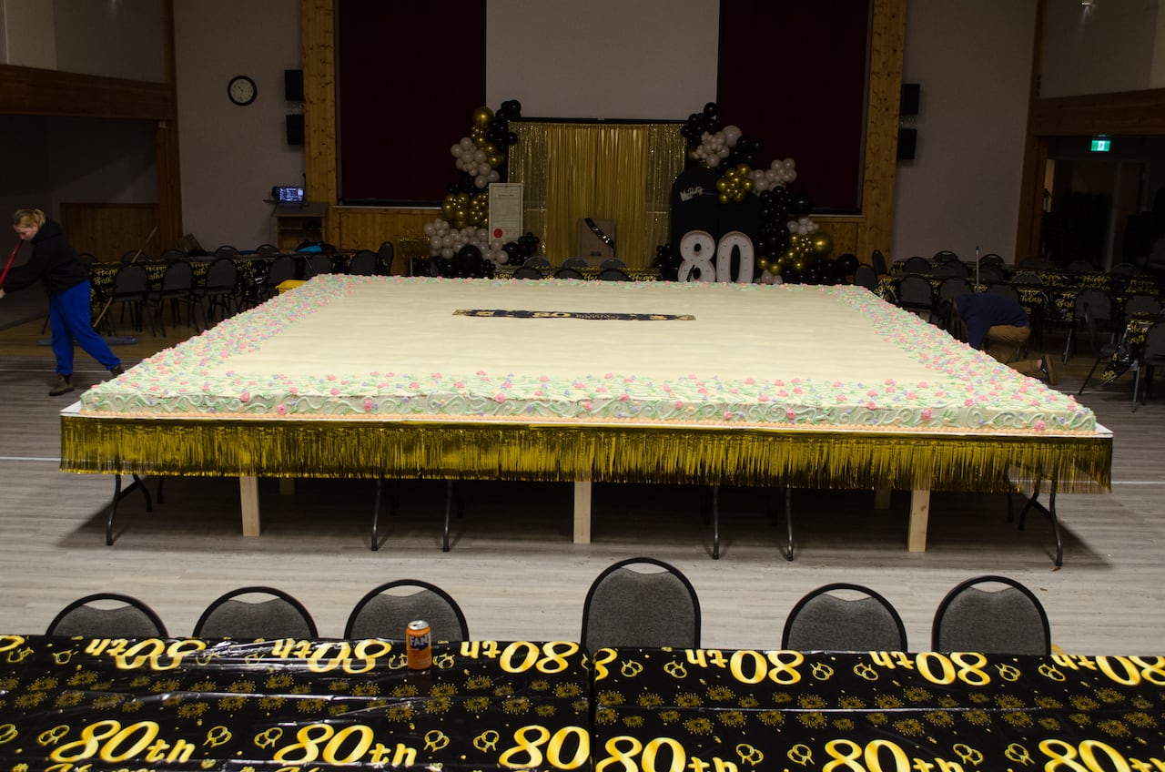 A massive cake sits in a community centre, with the numerals '80' on decorations throughout the room.