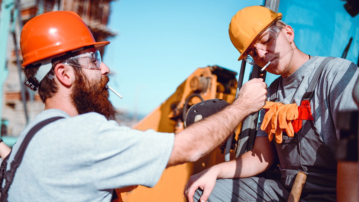 Male Construction Worker And Bulldozer Driver Taking A Cigarette Break outside, one lighting the other's cigarette