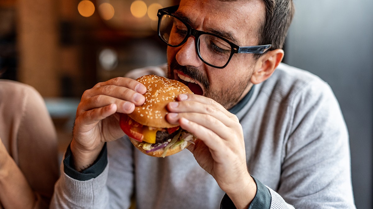 Man with glasses biting into a burger at a restaurant.
