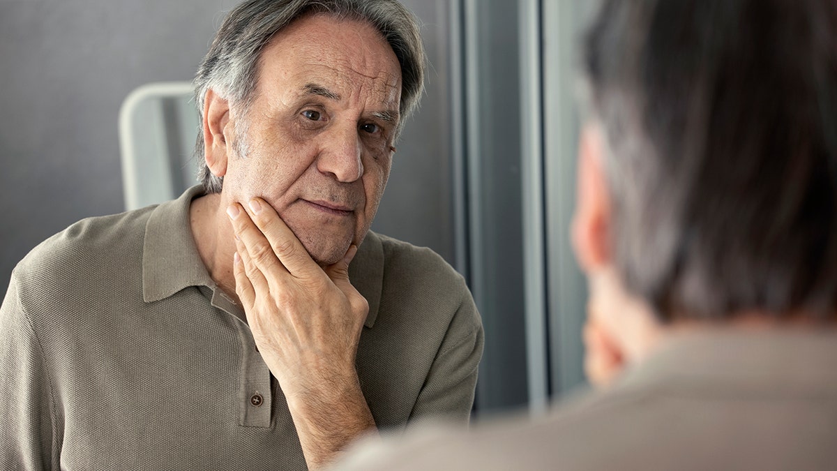 Older man with gray hair looking in mirror at his skin