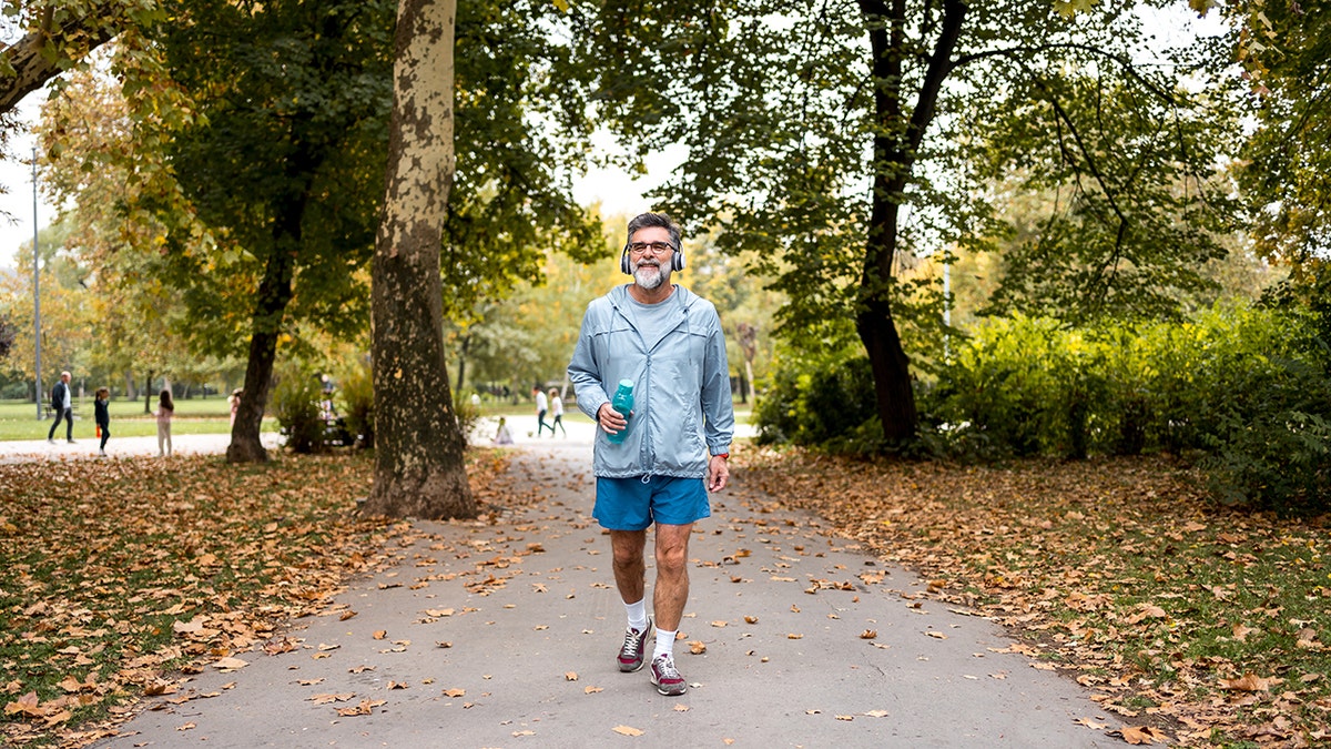 An older man walks along a park path with headphones on and a water bottle in hand.
