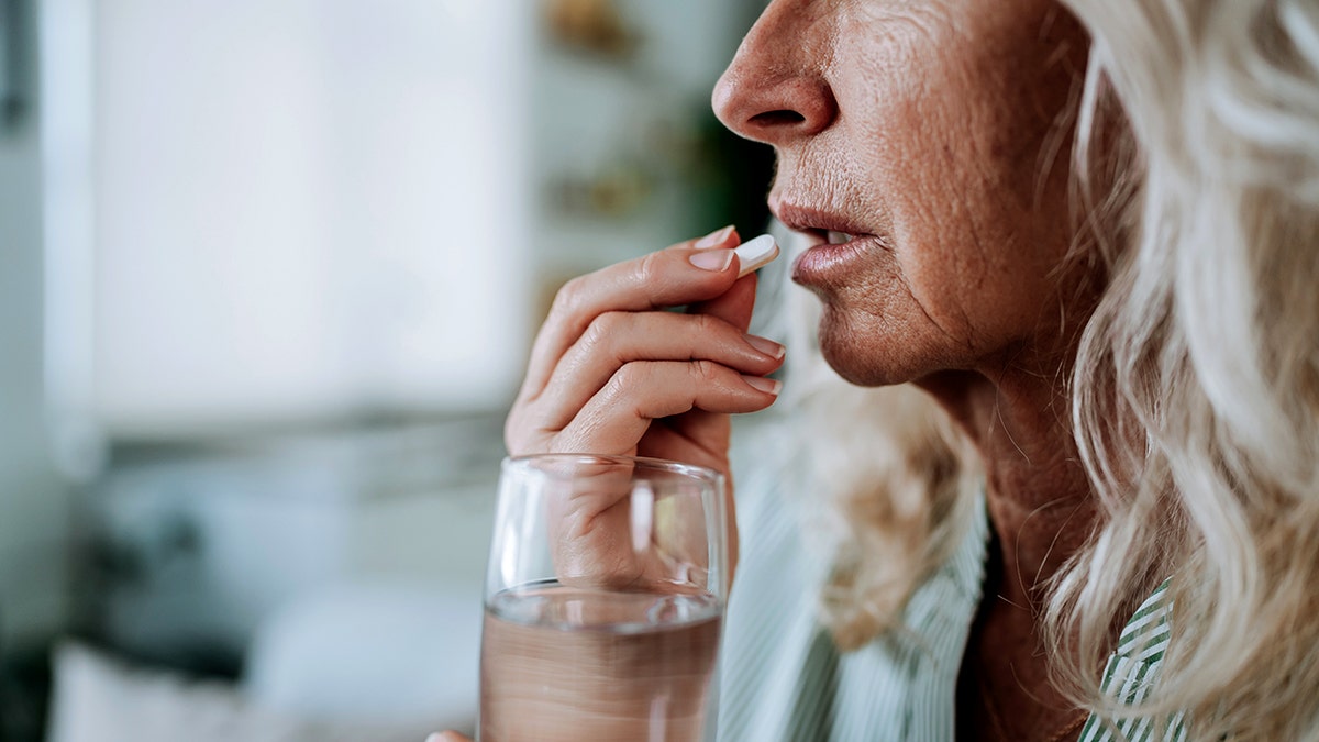 Profile view of a mature woman holding a white pill near her mouth, ready to take it with a glass of water.