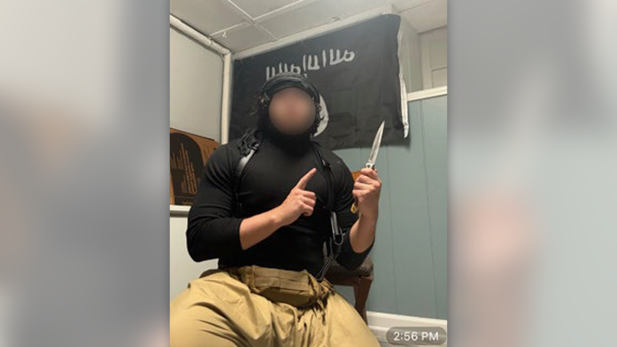 An image of a man whose face has been blurred holding a knife and posing in front of an ISIS flag