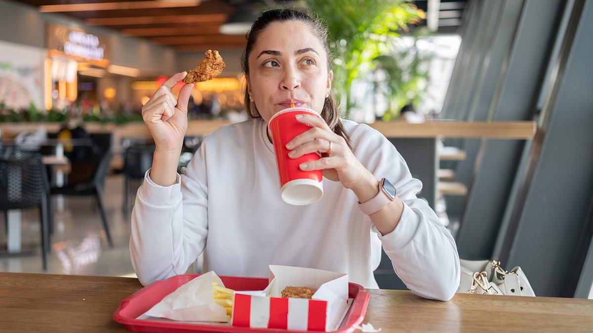 Woman drinking soda and holding a fried chicken wing while eating a fast food meal at a modern food court.