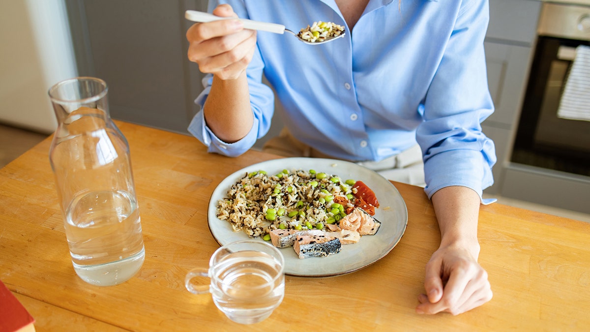 Person eating a healthy meal of salmon, rice, and vegetables at a wooden table.