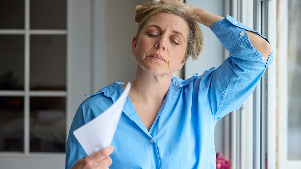 A menopausal woman cools herself at home during a hot flash by fanning with papers.