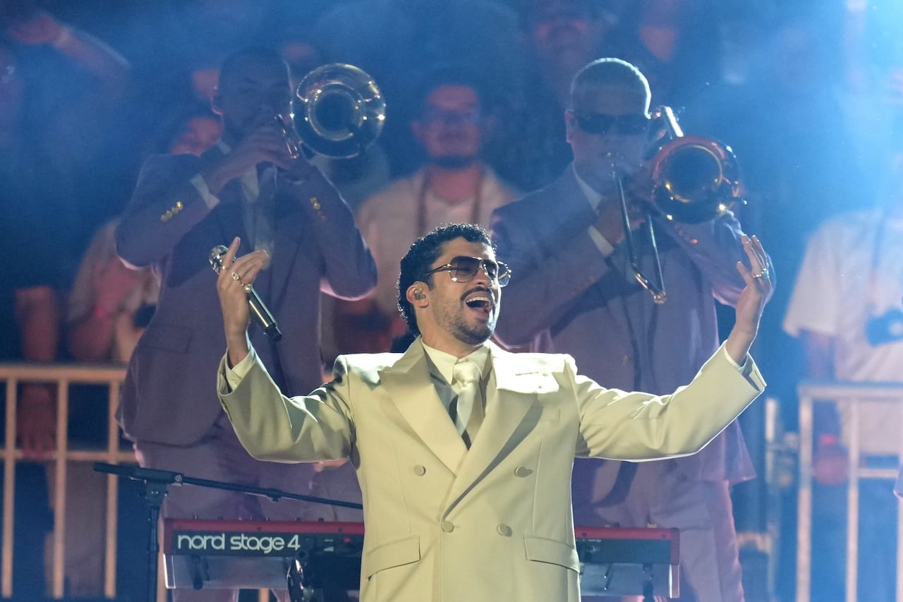A man in a tan suit and sunglasses holding a microphone, pictured from the waist up. Two people play trombones behind him.