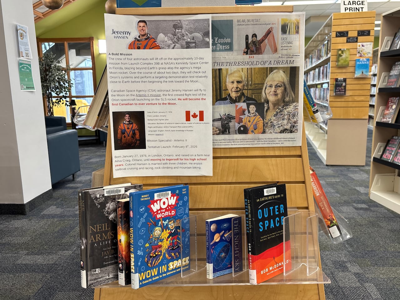 A library display featuring a poster of astronaut Jeremy Hansen, newspaper clipping and several space-themed books arranged on a wooden stand inside the Ingersoll Public Library.
