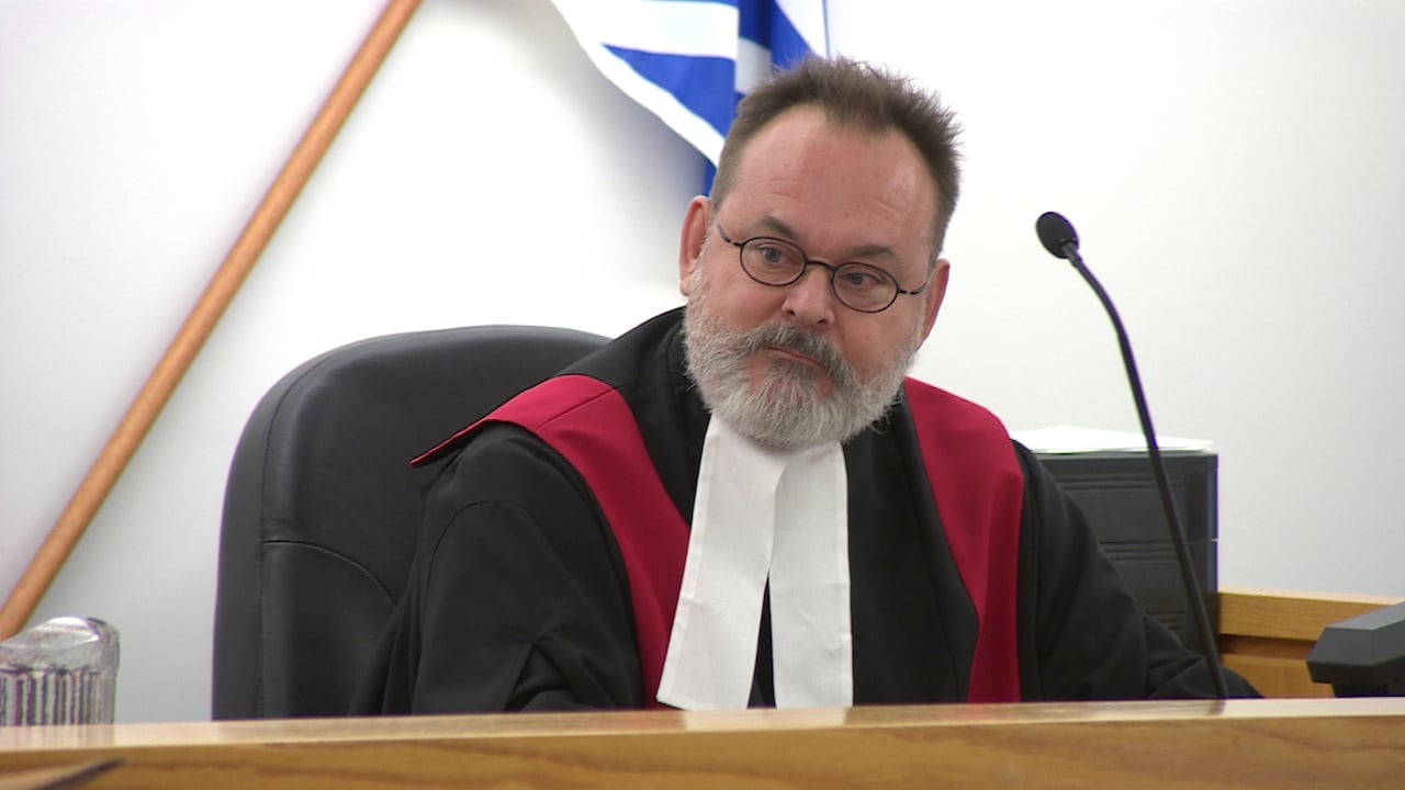 A man with a grey beard, glasses and wearing black and white robes sits at a bench with a microphone in front of him.