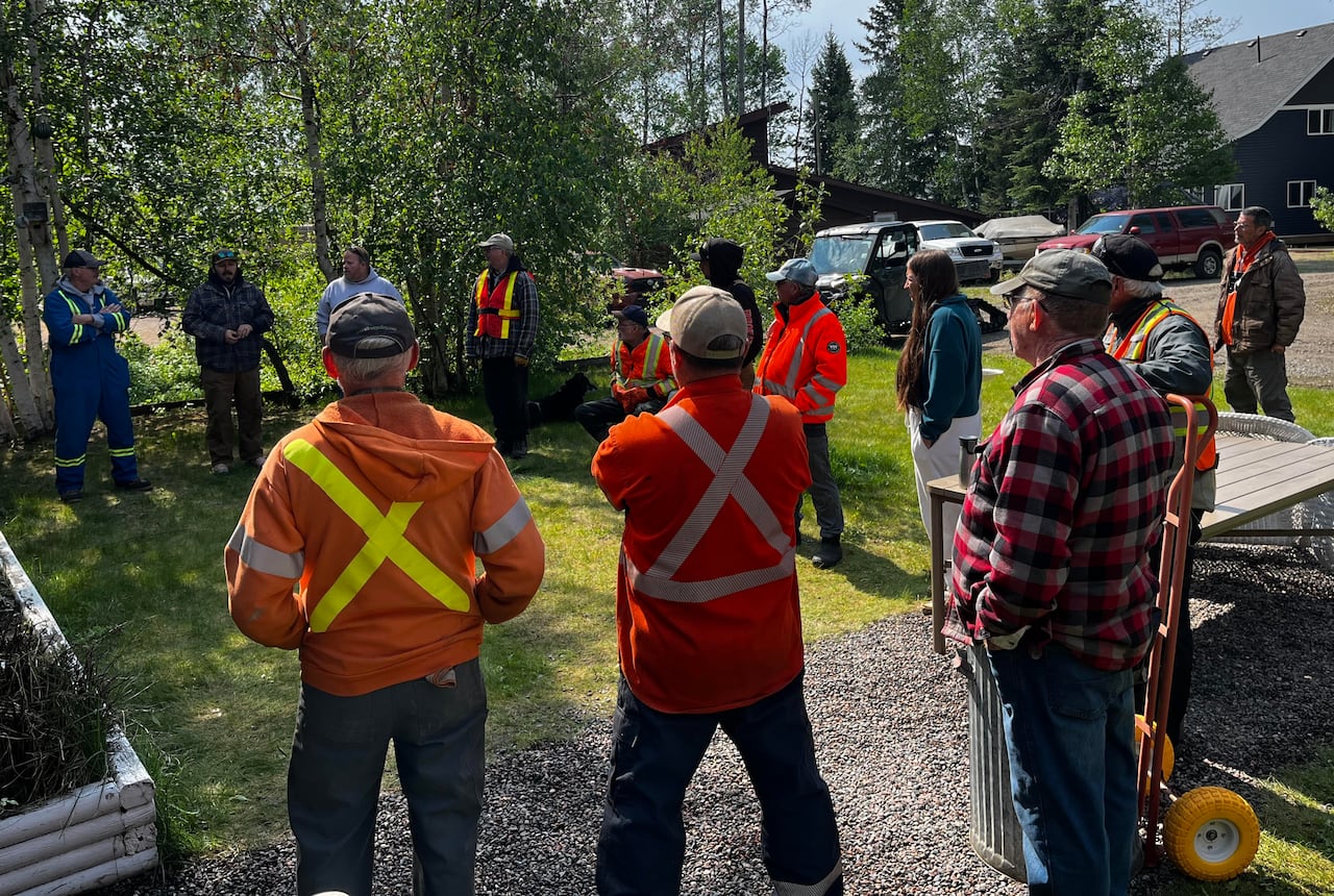 A group of people in orange vests and plaid jackets stand in a circle outside in the summer.