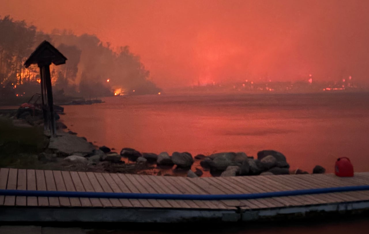 A lakeside dock with smoky red skies reflected in the water and fires in the background.