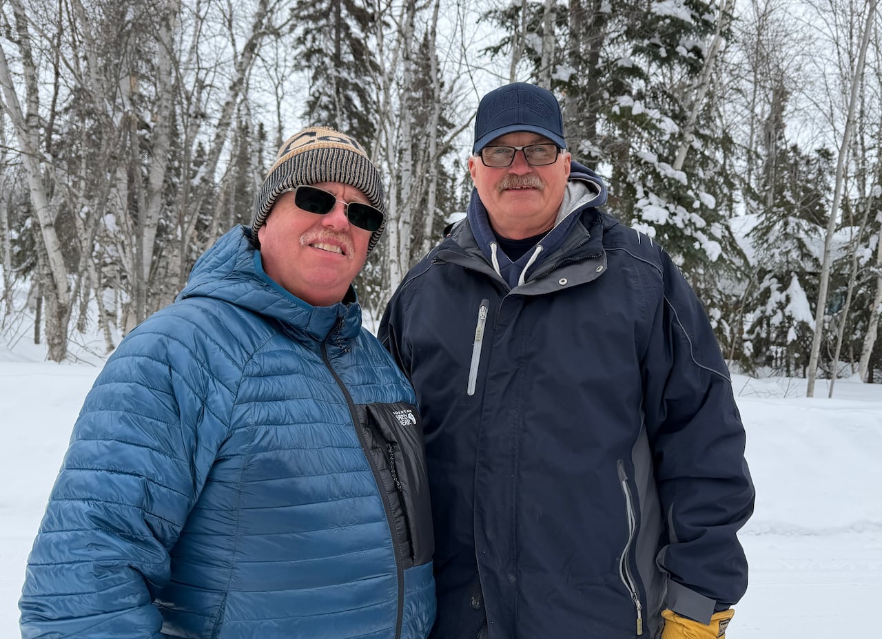 Two men in winter jackets stand in the middle of a snowy field, backed by trees.