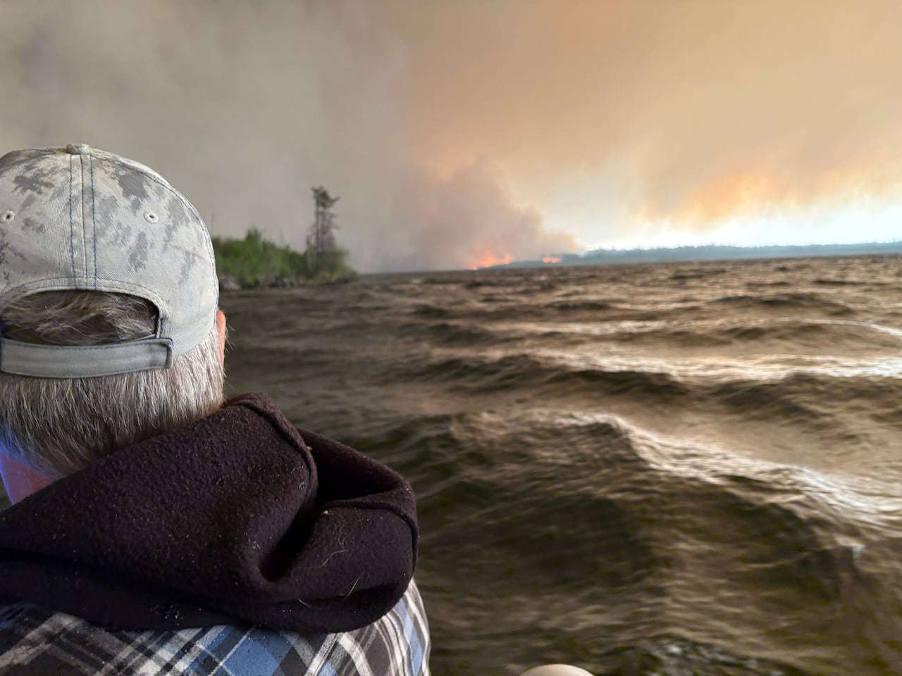A closeup of the back of a person's head shows them on the water, looking out at fires on the shore.