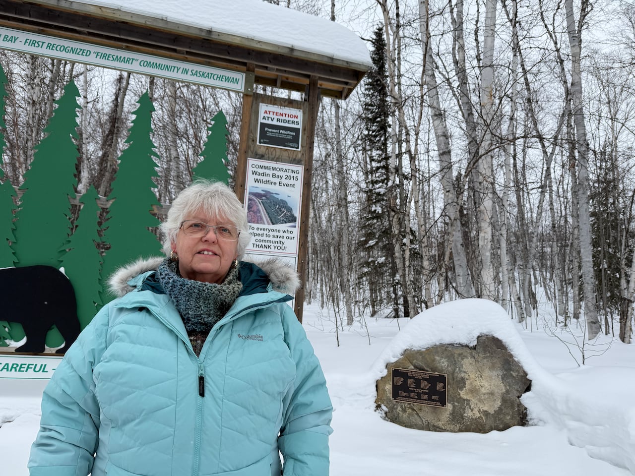 A white-haired woman in a blue winter jacket stands in the snow next to a rock with a plaque attached to it.