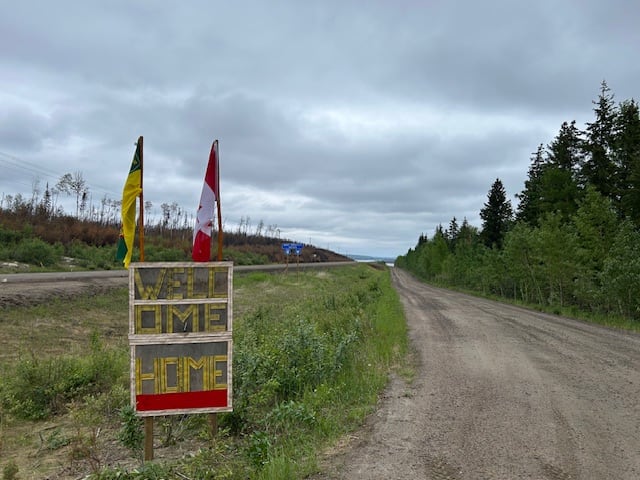 A sign reading 'Welcome home' stands beside a road, a Saskatchewan flag and a Canada flag mounted atop it.
