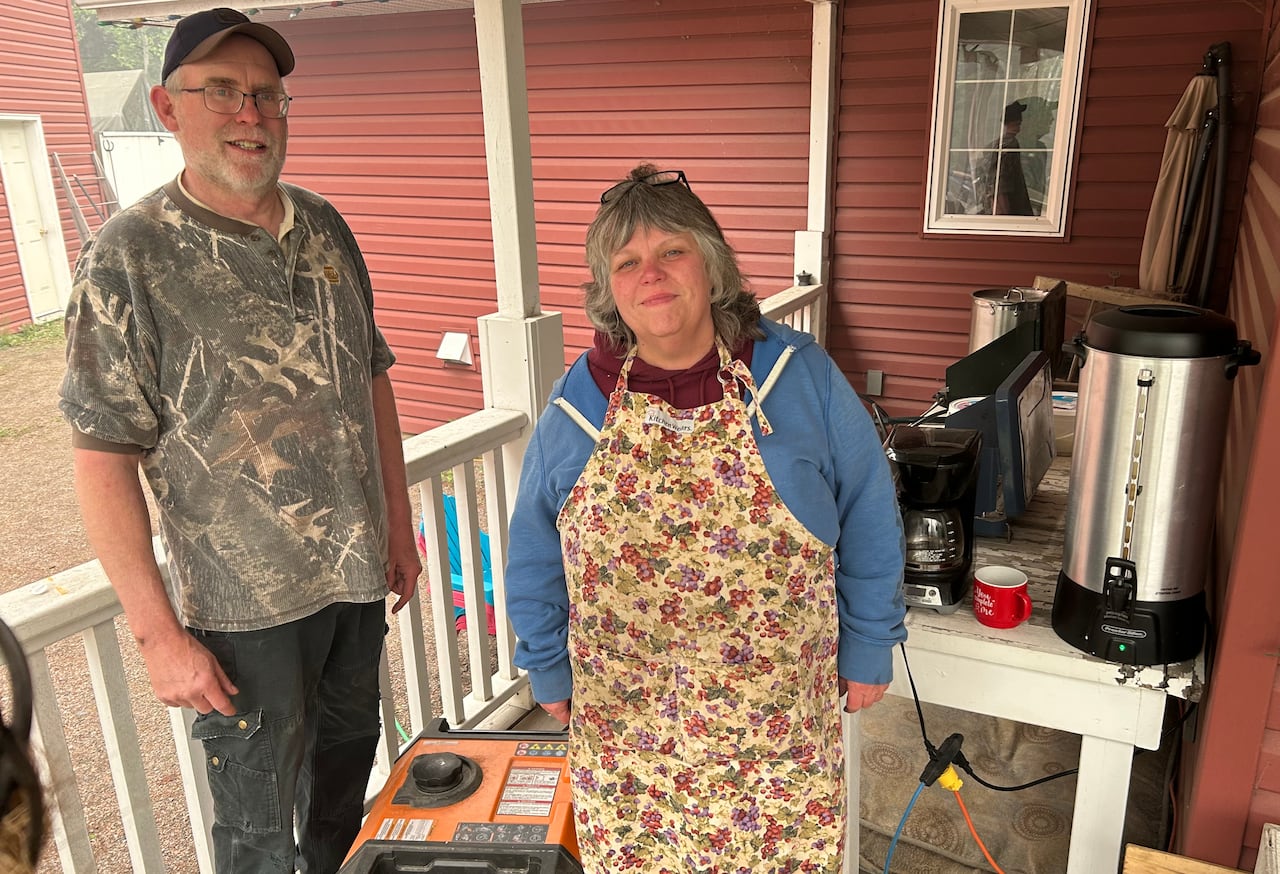 Two people stand on a deck outdoors, with a large coffee urn beside them.