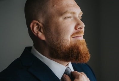 A bearded man in a dark suit adjusts his tie