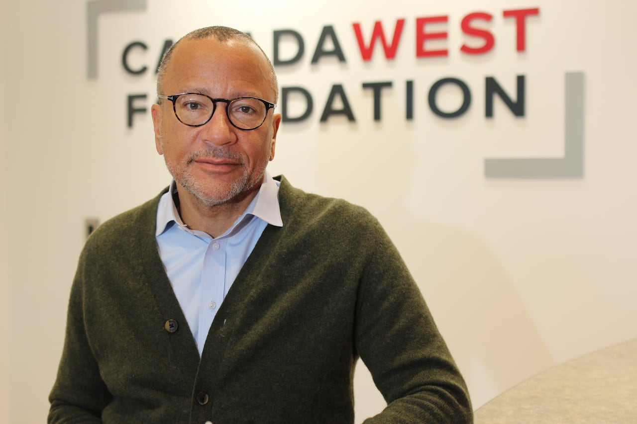 A man wearing glasses, a blue button-down shirt and a green cardigan poses in front of a wall bearing the logo of the Canada West Foundation.