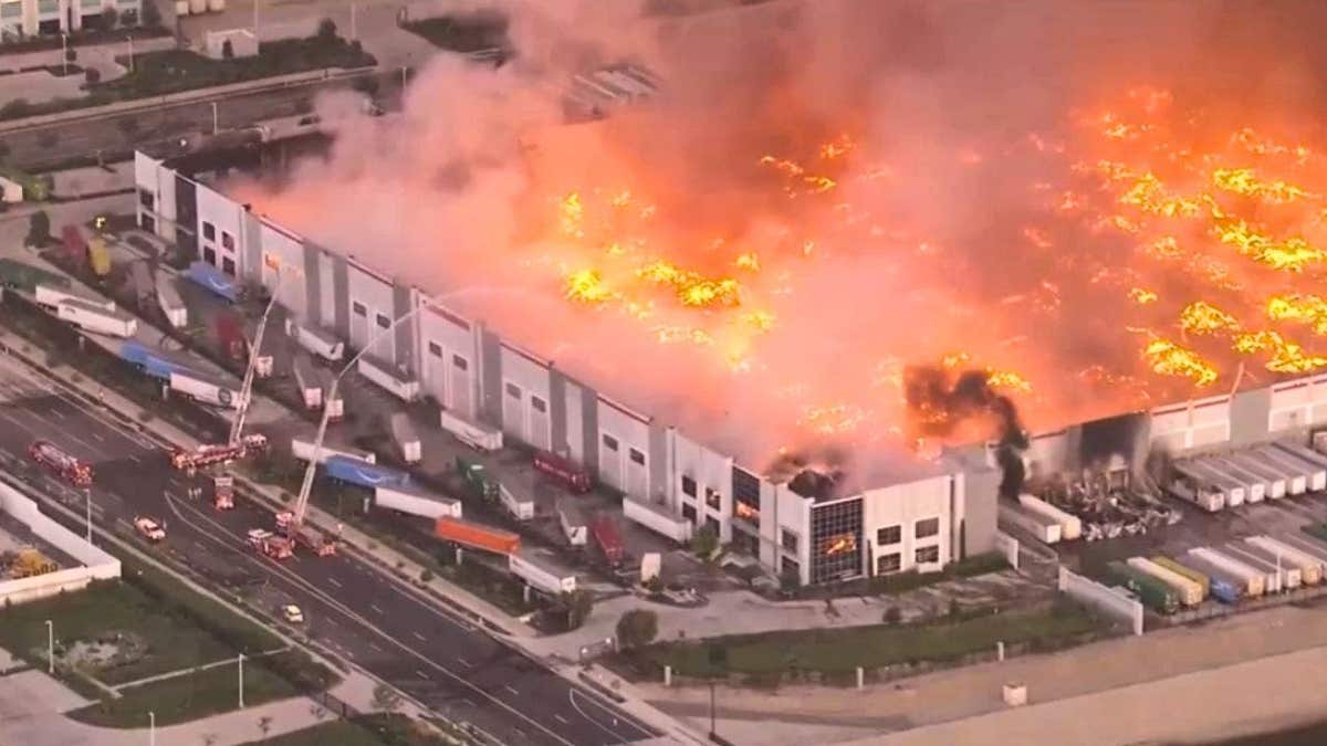 Aerial daytime image of a large warehouse on fire with flames and heavy smoke, as firefighters respond from the ground.