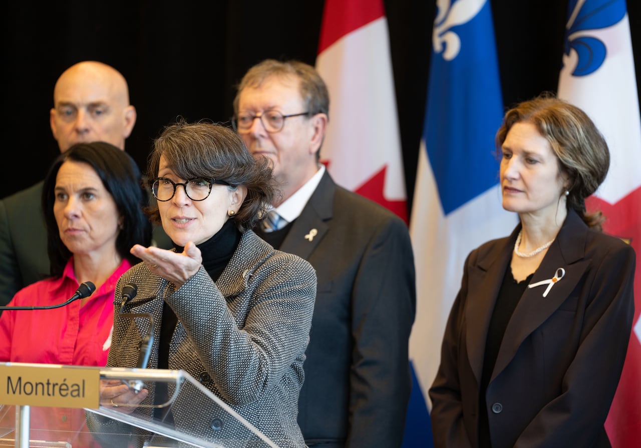 A woman speaks at a podium with two women, two men and large flags standing behind her.