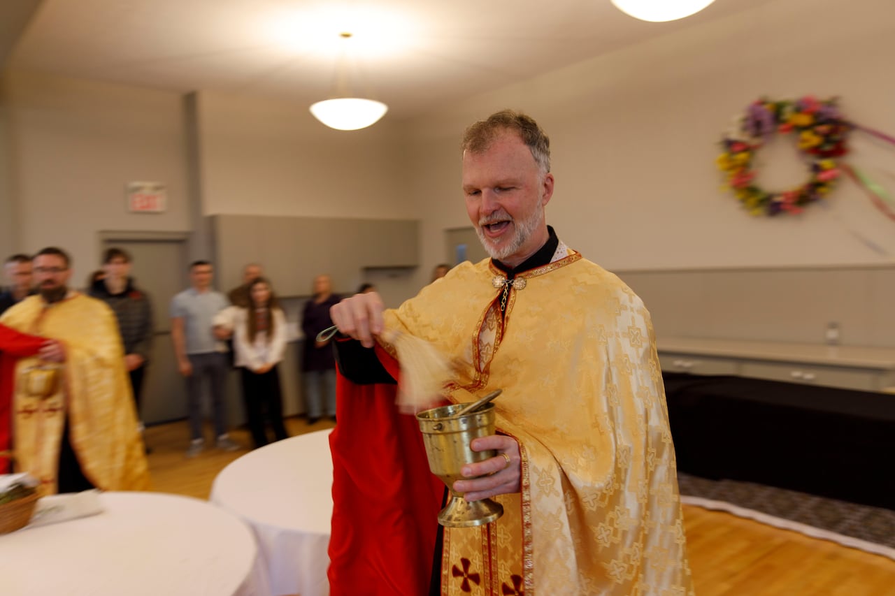 A father in robe holding a brass container and brush, with onlookers in the distance inside a church room.