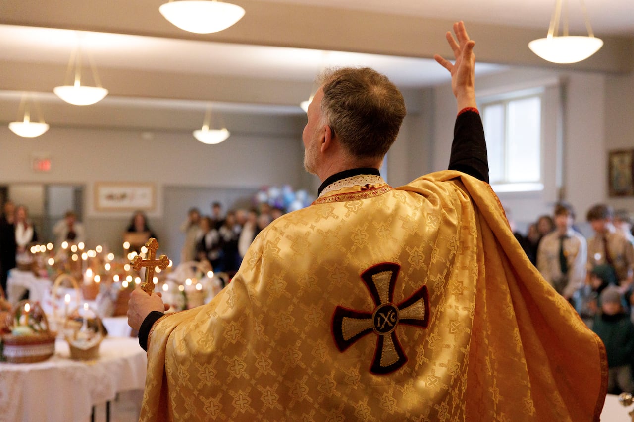 A father in robe holding a cross with one hand and gesturing with his other hand in front of churchgoers standing around tables filled with food baskets.