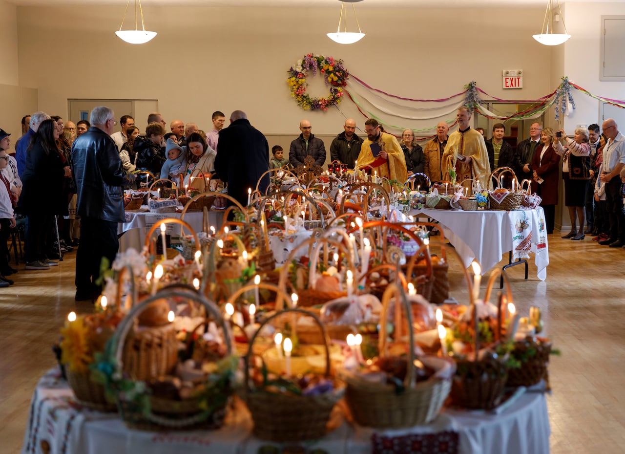 Food baskets with candles lit laid out on white-cloth tables, with people standing in the back of a church room.