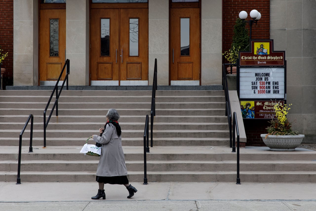 A woman in grey coat, carrying a basket and walking by the front of a church.