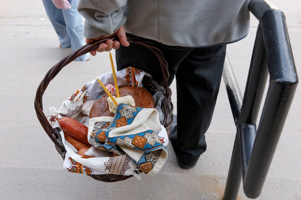 A closeup of the back of a person holding a basket in one hand filled with bread, sausages and candles, while the other hand holds a staircase railing and walking down steps.