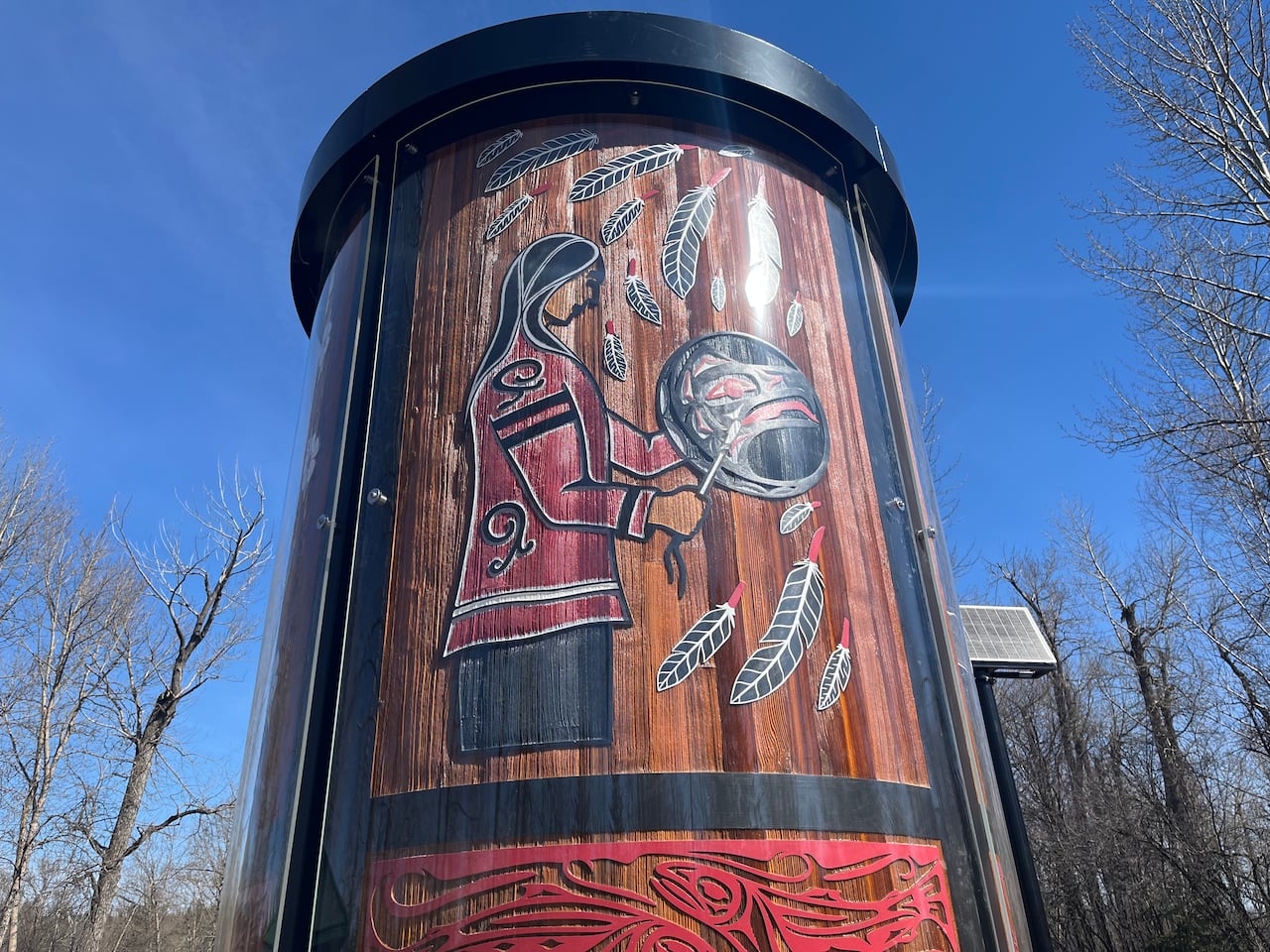 A cedar pillar depicts an Indigenous woman beating a drum surrounded by feathers. 
