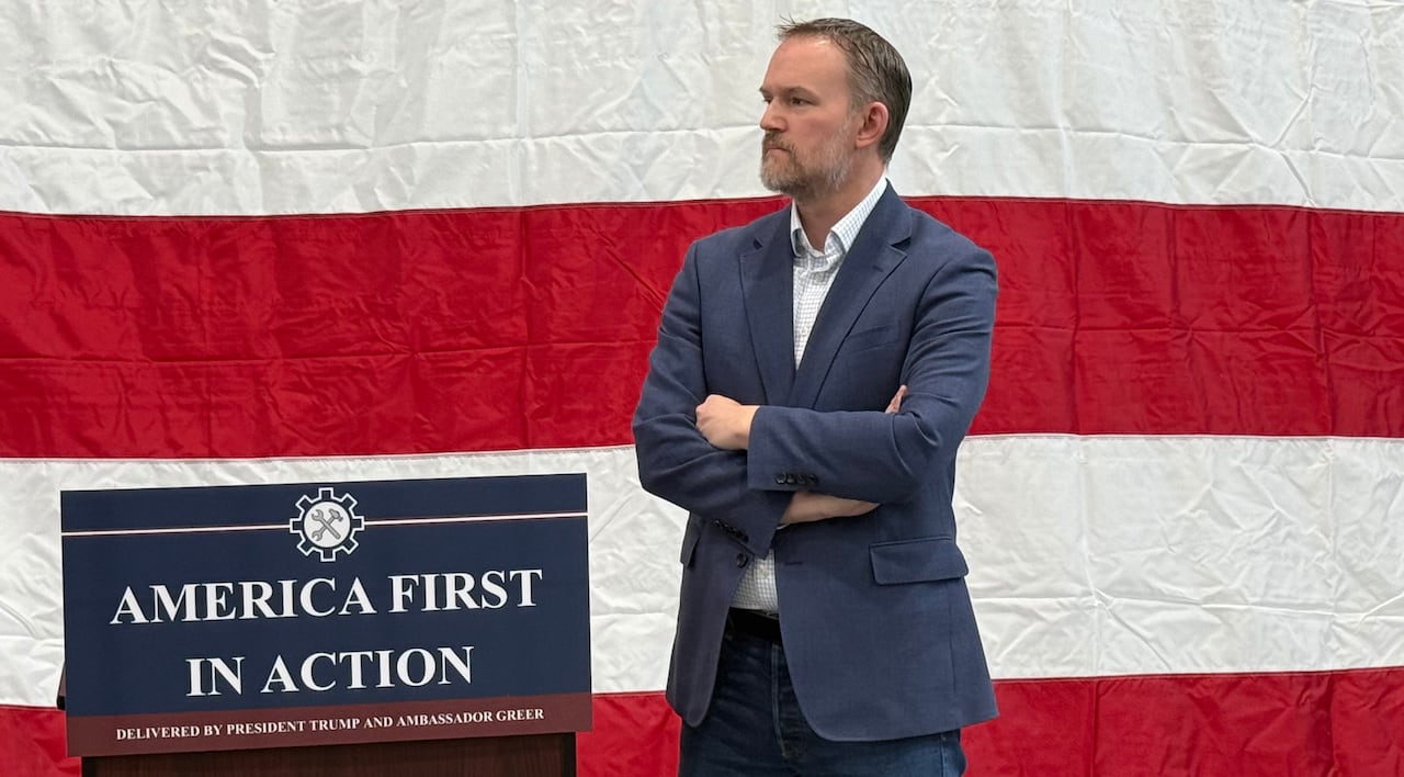 Jamieson Greer stands with his arms crossed in front of a backdrop of a large U.S. flag, beside a podium with a sign saying "America First in Action'