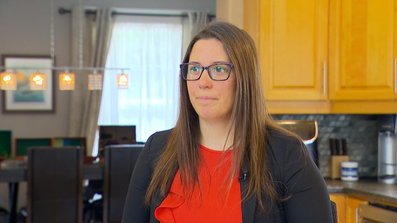 A woman with brown hair and glasses wears a red shirt and black blazer. She sits in her kitchen.