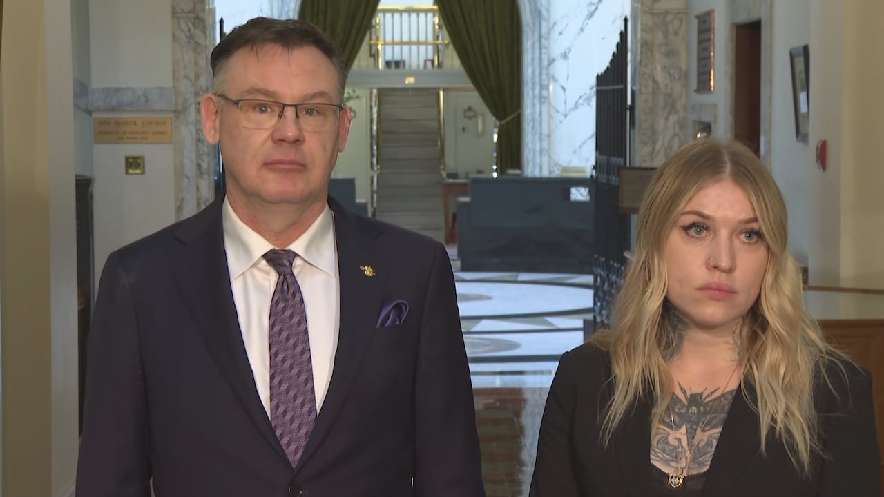 A tall white man and a woman with tattoos speak in a hallway.