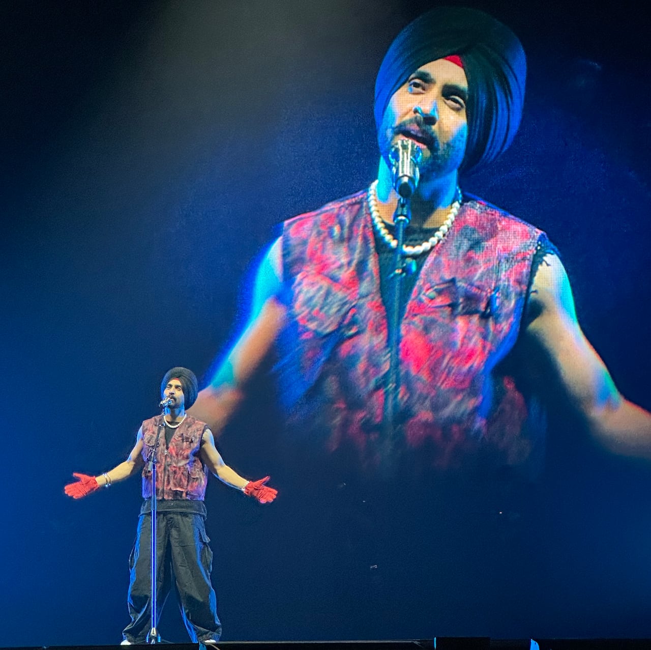 A performer sings into a microphone with outstretched arms. He is also shown on a video screen in the background.