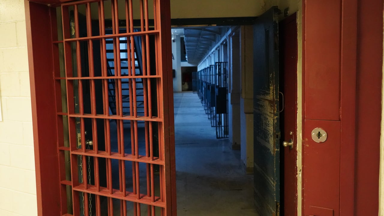 A row of open cell doors are shown in a dark hallway behind a set of red-painted bars.