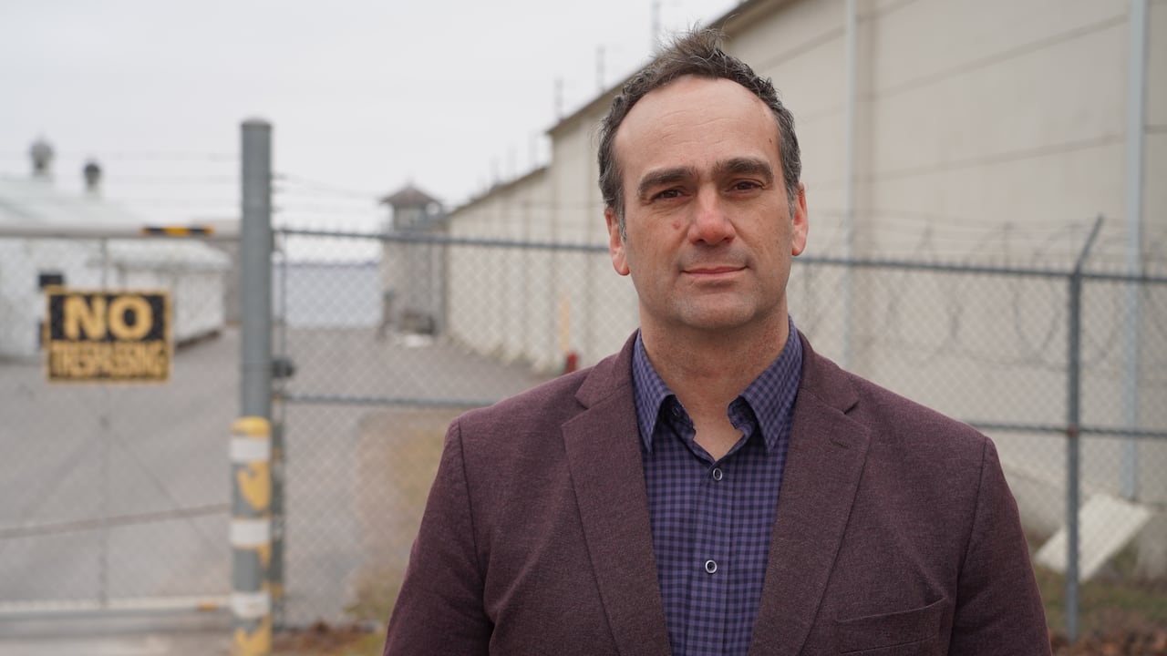 A man in a burgundy suit jacket stands next to a prison wall and guard tower on a cloudy day.